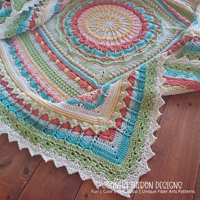 May include: A colorful, handmade crochet blanket with a circular design. The blanket features a variety of colors, including pink, green, blue, and yellow. The edges are scalloped. The text "JOANITA THERON DESIGNS" is visible at the bottom.