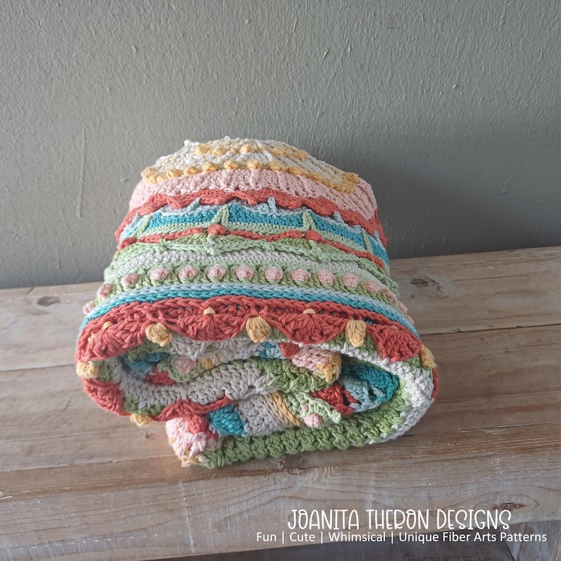May include: A folded, colorful crochet blanket with a variety of textures and patterns. The blanket features bands of pink, blue, green, and orange, with decorative elements. The text "JOANITA THERON DESIGNS" is visible at the bottom, along with the phrases "Fun | Cute | Whimsical | Unique Fiber Arts Patterns."