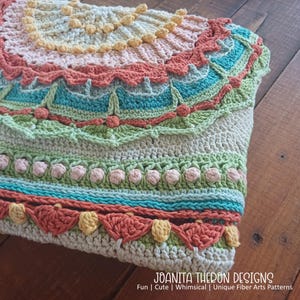 May include: A handmade crochet item in a variety of colors, including peach, teal, green, and cream. The design features intricate patterns and textures, with scalloped edges and floral accents. The text "JOANITA THERON DESIGNS" is visible at the bottom.