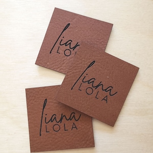 Puede incluir: Tres etiquetas de cuero marrón con el texto "liana LOLA" en letras negras.