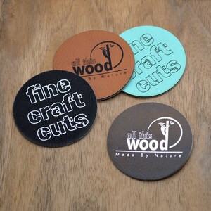 Puede incluir: Cuatro posavasos redondos con diferentes diseños. Los posavasos están hechos de cuero marrón y tienen texto blanco. El texto en los posavasos dice "all this wood", "fine craft cuts", "all this wood", y "the craft cuts".