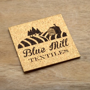 May include: A cork coaster with a black and white design of a farmhouse, trees, and rolling hills. The text "Blue Mill Textiles" is printed below the design.