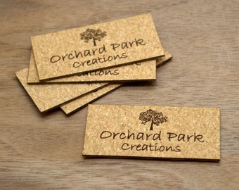 May include: Light brown rectangular cork name tags, featuring the text "Orchard Park Creations" and a tree graphic. The tags are arranged on a wooden surface.
