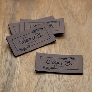 May include: A collection of brown rectangular labels with the text "Naomi B. SEWIST" in a handwritten font, framed by a thin black border and decorative leaves. The labels are arranged on a wooden surface.
