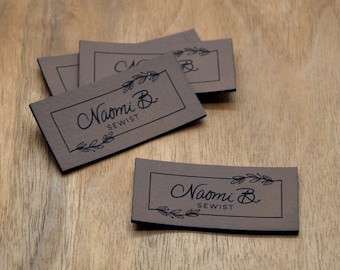 May include: A collection of brown rectangular labels with the text "Naomi B. SEWIST" in a handwritten font, framed by a thin black border and decorative leaves. The labels are arranged on a wooden surface.