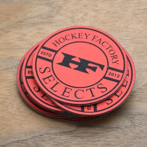 Puede incluir: Una pila de posavasos redondos rojos con texto negro. Los posavasos tienen las palabras "HOCKEY FACTORY SELECTS" en un diseño circular. El centro del posavasos tiene las letras "HF" en negro. Sobre una superficie de madera.