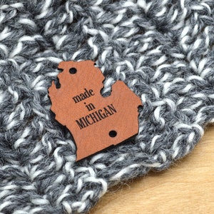May include: Close-up of a gray and white knitted fabric with a brown leather tag. The tag is shaped like the state of Michigan and has the words "made in MICHIGAN" printed on it. The knitted fabric appears to be a scarf or hat.