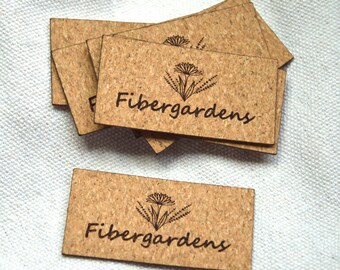 May include: A collection of rectangular cork tags, each displaying the text "Fibergardens" in a handwritten style, alongside a floral emblem. The tags are a light brown hue, suggesting a cork material.