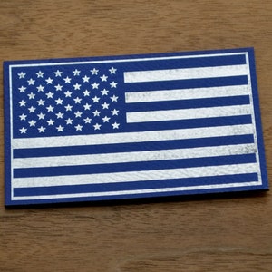 Puede incluir: Una tarjeta rectangular con el diseño de la bandera estadounidense. La bandera tiene un campo azul con estrellas blancas y rayas blancas y azules alternas. La tarjeta está sobre un fondo de madera.