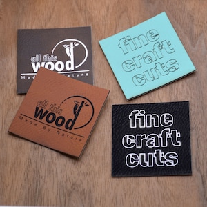 May include: Four square leather labels with different designs. Two labels are brown with the text "all this wood made by nature" and a bird illustration. One label is turquoise with the text "fine craft cuts" in white. One label is black with the text "fine craft cuts" in white.