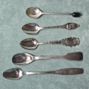 May include: A collection of five silver-toned metal spoons arranged vertically. The top spoon has a black bead handle. The second features a New Mexico engraving and a figure. The third has an ornate handle. The bottom two have plain handles.