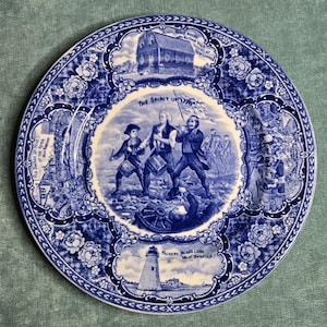 May include: Blue and white decorative plate featuring historical scenes and text. The central image depicts figures with a flag, labeled "The Spirit of 1776." Other scenes include a lighthouse, a schoolhouse, and text around the rim. Floral and scrollwork border.