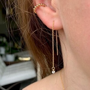 Puede incluir: Un pendiente de cadena de oro con un pequeño diamante en el extremo. El pendiente cuelga del lóbulo de la oreja y se combina con un ear cuff de oro.