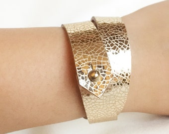 Könnte beinhalten: Ein goldfarbenes Lederarmband zum Umwickeln mit strukturierter Oberfläche. Das Armband hat einen kleinen goldfarbenen Knopfverschluss.