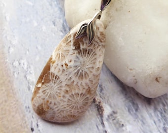 May include: A teardrop-shaped pendant necklace featuring a fossilized coral stone with a tan and white starburst pattern. The stone is set in a polished silver-toned metal bail and hangs from a matching silver-toned metal chain link necklace.