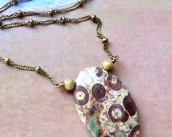 May include: An oval-shaped pendant featuring a mottled pattern of dark brown circles on a cream and sage green background. The pendant is suspended from a metallic bronze-toned chain that incorporates small, evenly spaced spherical beads.