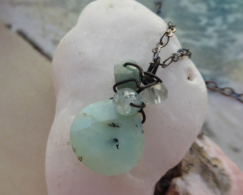 May include: A delicate necklace with a cluster of light blue and clear gemstones wrapped in wire. The necklace is made with a dark metal chain.