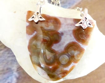 May include: A pendant necklace featuring a polished, shield-shaped piece of orbicular jasper with brown, tan, and cream circular patterns. The stone is suspended from a silver-toned metal chain by two decorative, triangular-shaped metal bails.