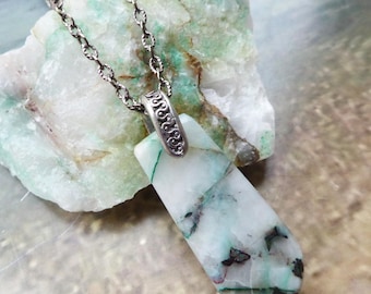 May include: A necklace featuring a polished, teardrop-shaped stone pendant with white, green, and black mineral inclusions. The pendant is attached to a silver-toned metal bail with an embossed scroll pattern, suspended from a matching silver-toned chain.