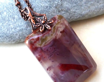 May include: A rectangular pendant necklace with a copper-colored chain. The pendant is a polished stone with a mix of red, purple, and white hues. The pendant's top has a decorative copper-colored floral design.