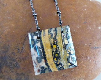 May include: A square pendant featuring a polished, multi-colored stone with swirling patterns of yellow, black, white, and grey, suspended from a dark metallic chain with small, evenly spaced beads along its length.