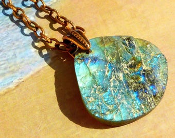 May include: A pendant necklace featuring a flat, irregular, polished gemstone with iridescent blue and green flashes. The stone is suspended from a textured, copper-toned metal chain that connects to a decorative, ridged metal bail at the top.