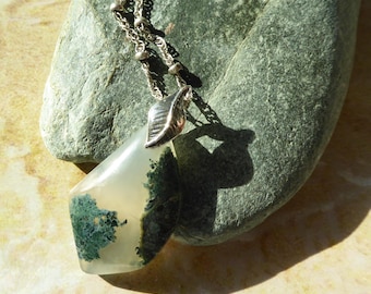 May include: A pendant necklace featuring a translucent, teardrop-shaped moss agate stone with green dendritic inclusions. The stone is held by a silver-toned metal bail shaped like a leaf, attached to a delicate silver-toned chain with small beads.