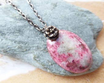 May include: A necklace featuring an oval-shaped pink and white mottled stone pendant suspended from a silver-toned chain. The pendant is topped with a small, detailed metal flower charm that connects the stone to the chain links.