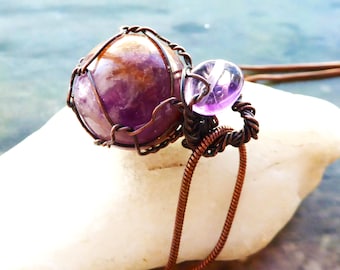 May include: A handcrafted pendant featuring a large, round amethyst gemstone encased in a copper wire cage. A smaller, teardrop-shaped amethyst bead is attached. The pendant hangs from a brown cord, resting on a white stone.