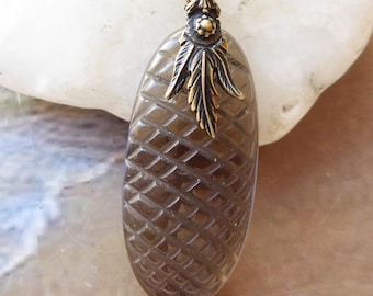 May include: An oval-shaped, translucent brown gemstone pendant featuring a carved diamond-grid pattern on its surface. The pendant is attached to a dark bronze-toned metal bail shaped like a leaf and hangs from a matching bronze-toned chain.