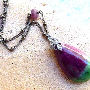 May include: A silver-toned necklace with a teardrop-shaped pendant. The pendant is a mix of deep purple and green, with a silver floral accent. The necklace chain has small beads and a delicate design, perfect for adding a touch of elegance.