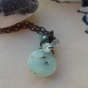 May include: A delicate necklace with a light blue, oval-shaped pendant and smaller light blue and clear gemstone accents. The necklace is made with a dark gray chain.