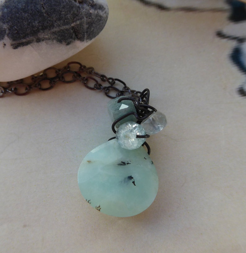 May include: A delicate necklace with a light blue, oval-shaped pendant and smaller light blue and clear gemstone accents. The necklace is made with a dark gray chain.