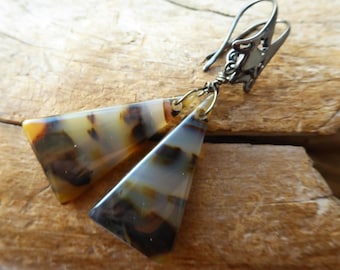 May include: A pair of dangle earrings featuring two triangular, translucent brown and cream mottled stone pendants suspended from dark metal ear wires. The pendants are attached to the wires with small metal jump rings and wire wrapping.