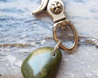 May include: A brass-toned metal swivel lobster clasp keychain featuring an ornate, embossed circular design on the central hinge. Attached to the bottom loop is a smooth, polished, olive-green stone pendant secured by a small silver-toned metal wire.