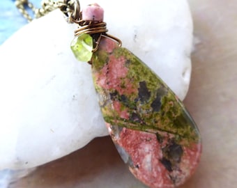 May include: A handcrafted pendant necklace featuring a teardrop-shaped unakite stone. The stone displays a mix of pink, green, and black hues. The pendant is accented with a small pink bead, a green gemstone, and copper wire. The necklace chain is a bronze color.