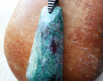 May include: A pendant featuring a trapezoidal stone with mottled green, black, and deep red coloration. The stone is held by a silver-toned metal bail with horizontal ridges, attached to a thick, braided silver-toned metal chain necklace.