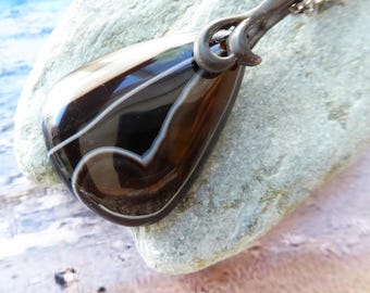 May include: A teardrop-shaped pendant necklace featuring a polished dark brown and black banded agate stone with distinct white concentric lines. The stone is encased in a dark, oxidized metal setting with a decorative loop at the top.