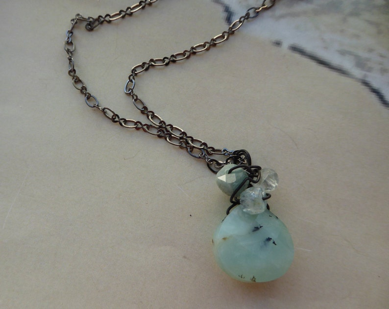May include: A silver chain necklace with a light blue and white gemstone pendant. The pendant is made of a large, teardrop-shaped stone with smaller stones clustered around it.