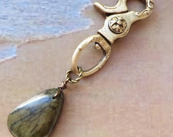 May include: A brass lobster claw clasp featuring an ornate circular detail with a floral pattern. Attached to the clasp is a teardrop-shaped stone pendant in shades of olive green and brown, secured by a twisted wire loop and a small metal ring.