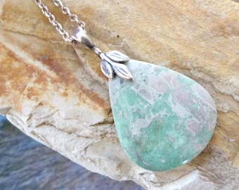 May include: A teardrop-shaped pendant necklace featuring a polished green and grey mottled stone. The stone is set in a silver-toned metal bail designed with three small leaf-like accents at the top, suspended from a matching silver-toned rope chain.