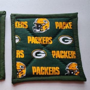 May include: Two green and gold Packers pot holders. The pot holders feature the team's logo, helmet, and the word "Packers" in gold lettering. The pot holders are square and have a green border.