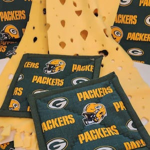 Op de afbeelding: Green Bay Packers-thema-artikelen. De afbeelding toont twee vierkante pannenlappen met een groene achtergrond en de woorden "PACKERS" en "KERS" in het geel. Op de achtergrond een gele kaasdoek met gaten.