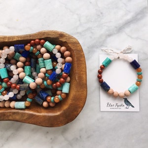 May include: A close-up of a wooden bowl filled with colourful gemstone bracelets. The bracelets are made with various stones, including blue lapis lazuli, green aventurine, red jasper, and brown wood beads. A single bracelet is displayed on a white card with the text "Blue Raven Handcrafted".