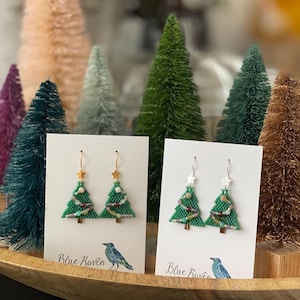 May include: Green beaded Christmas tree earrings with gold star accents. The earrings are displayed on white cards with the text "Blue Raven" and a bird illustration. Miniature Christmas trees are in the background.