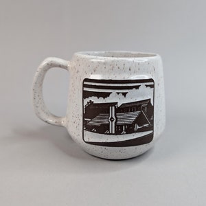 May include: White ceramic mug with a brown speckled glaze. The mug features a brown graphic of a building with a roof and a cloudy sky.