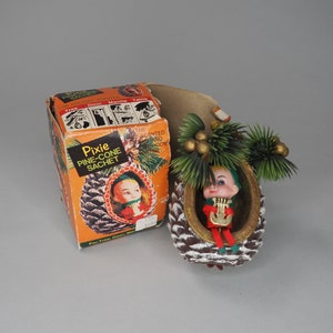 May include: A vintage Christmas bauble shaped like a pine cone with a small elf figurine inside. The elf is wearing a red and green outfit and is holding a musical instrument. The bauble is packaged in a box with the text "Pixie Pine-Cone Sachet" on it.