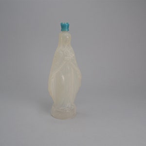 May include: A translucent, Madonna-shaped bottle with a light blue crown-like stopper. The bottle has a detailed design of a draped figure. The bottle is standing upright against a plain, light grey background.