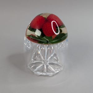 May include: A clear bottle stopper with a dome containing red berries and green leaves. The stopper is set on a clear, faceted base, creating a unique and elegant design for a bottle.
