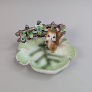 May include: A ceramic squirrel figurine on a leaf-shaped dish. The squirrel is brown and white with a smiling face. A small brown fence with green and blue floral accents sits behind the squirrel. The dish is light green.